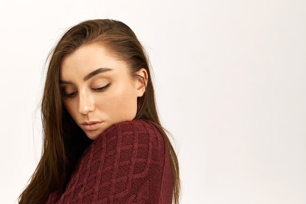 Beauty, style and fashion concept. Studio shot of shy beautiful young woman with perfect shiny skin posing isolated in warm sweater, being cold, having timid facial expression, looking down