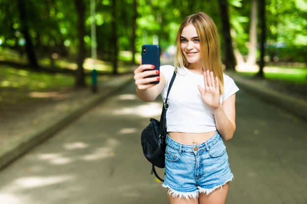 晴れた日に屋外の公園でスマートフォンでselfieを取っている10代の美少女。