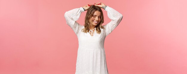 Beauty tenderness and fashion concept Attractive blond caucasian woman with tattoos in light white spring dress raise hands up relaxed smiling with closed eyes dancing pink background