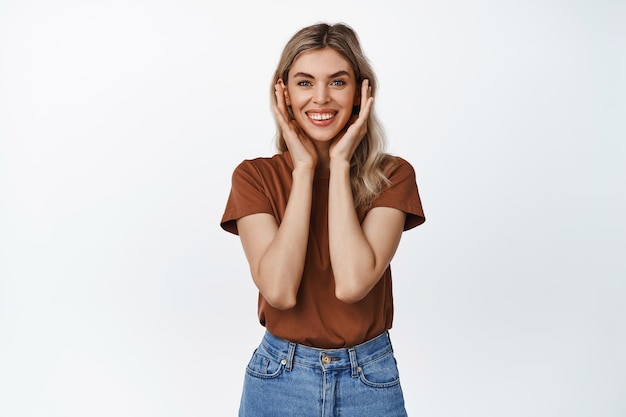 Beauty and wellbeing Smiling blond girl looking at camera holding hands on face standing in brown tshirt and jeans over white background