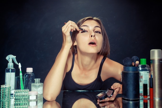 Donna di bellezza che applica trucco. bella ragazza guardarsi allo specchio e applicare cosmetici con un pennello. mattina, trucco e concetto di emozioni umane