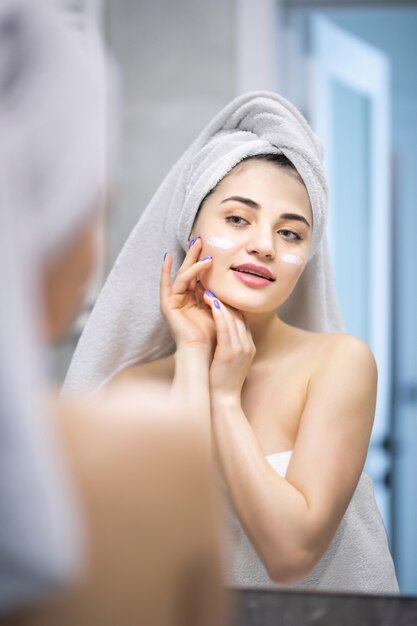 Beauty woman looking in mirror, applying hydrating lotion creme on cheeks, finishing morning domestic skincare routine