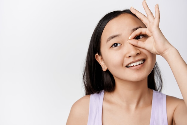 Beauty. Young asian woman with clean glowing skin, showing ok, zero gesture on eye, smiling and gazing away, standing on white.