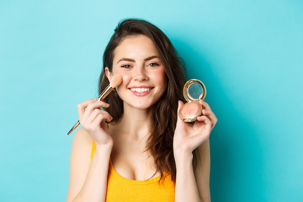 Beauty. Young attractive woman smiling, applying make up with brush, showing blushes at camera, standing over blue background