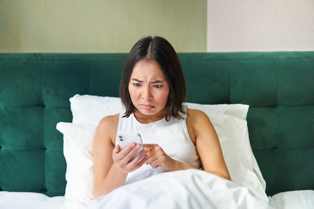 Бесплатное фото Фотография спальни азиатской женщины, лежащей в постели, выглядящей испуганной и шокированной чтением с экрана смартфона