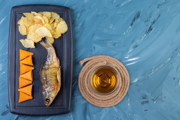 파란색 배경에 있는 쟁반에 있는 칩과 말린 생선 옆에 있는 삼발이에 있는 맥주 유리.