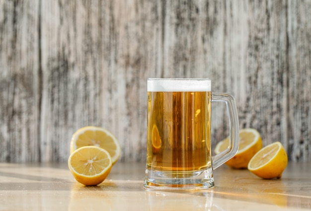 Пиво с дольками лимона в стеклянной кружке на шероховатый и светлый стол, вид сбоку.