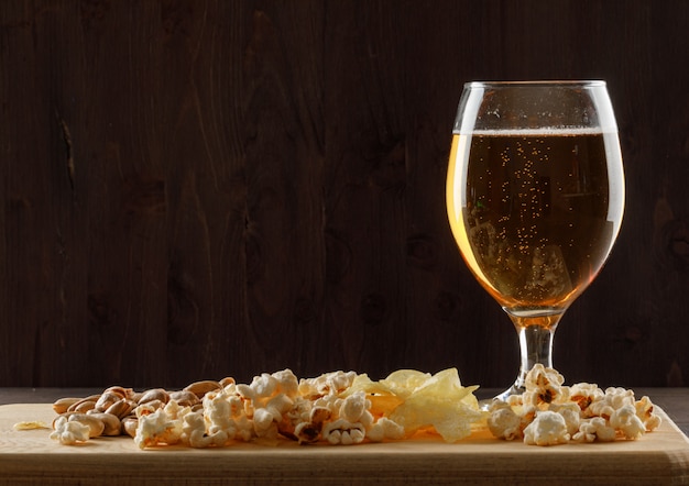 Beer with snack in a goblet glass on wooden and cutting board table, side view.