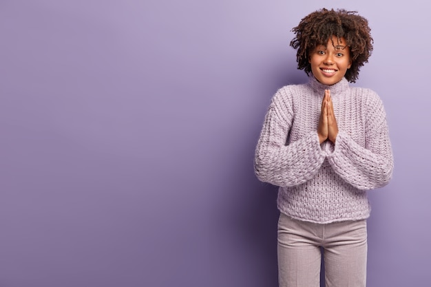 物乞いをする素敵なアフリカ系アメリカ人の女性は、陽気な表情を求め、彼女をサポートするように頼み、手のひらを祈りのジェスチャーで保ち、紫色の壁に隔離されたカジュアルな冬のセーターを着ています。