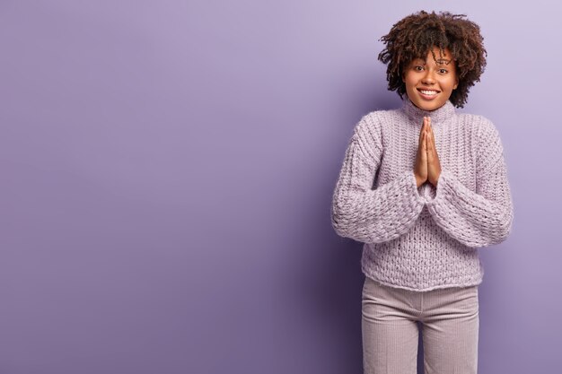 Begging lovely Afro American lady has beseeching cheerful look, asks to support her, keeps palms in praying gesture, wears casual winter sweater, isolated over purple wall.