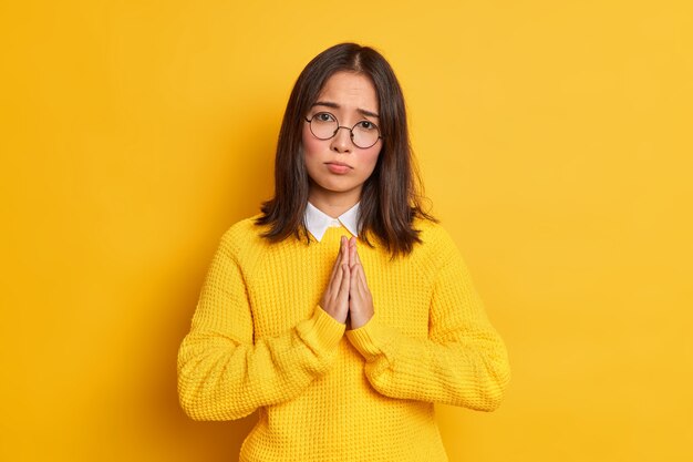 Begging pleading young Asian woman with dark hair keeps hands in pray gesture looks with imploring expression wears round spectacles and casual sweater.