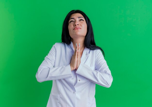 Begging young female doctor wearing medical robe looking up keeping hands together in pray gesture 