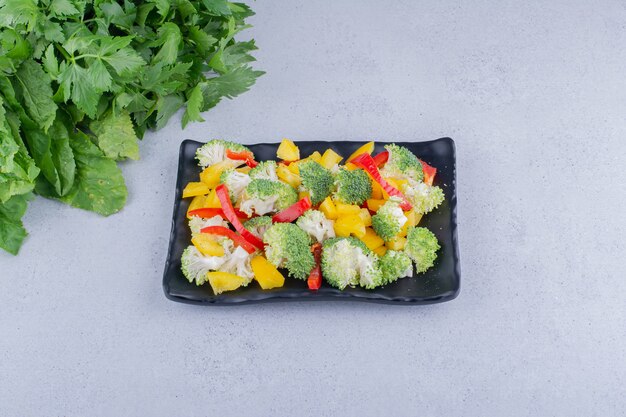 Bell pepper and broccoli salad on a platter next to a pile of greens on marble background. High quality photo