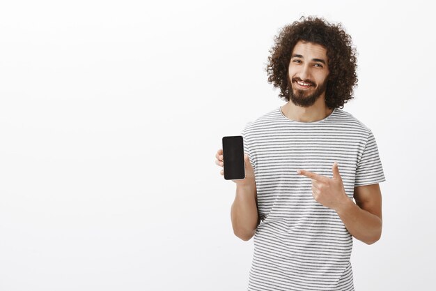 Best choice for modern people. Joyful handsome male model with beard in striped t-shirt, showing smartphone and pointing at device with index finger, smiling broadly