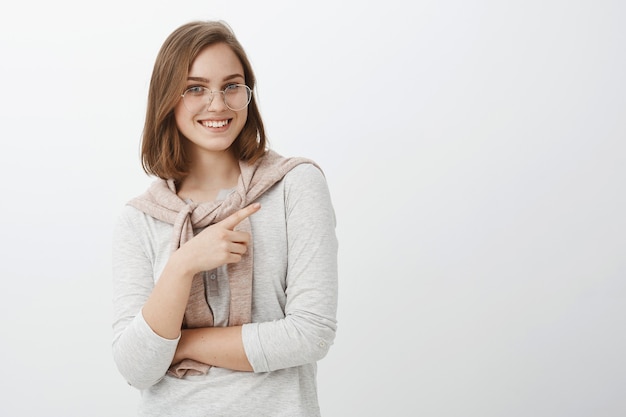 Best copy space you ever seen. Happy carefree beautiful young woman with brown short hair in eyeglasses and pullover tied over neck smiling friendly pointing right directing at cool place