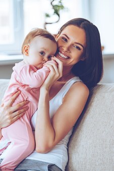 史上最高の抱擁。女の赤ちゃんを手に持って、自宅のソファに座って笑顔でカメラを見ている陽気な美しい若い女性