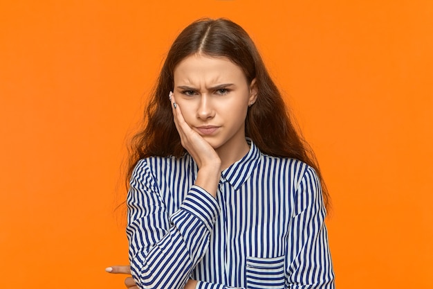 Beuatiful unhappy young Caucasian female wearing stylish striped blouse suffering from toothache