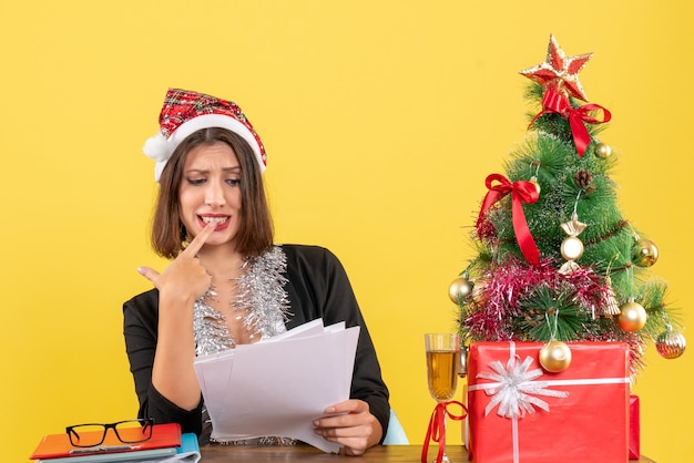 Bewildered business lady in suit with santa claus hat and new year decorations holding documets and sitting at a table with a xsmas tree on it in the office