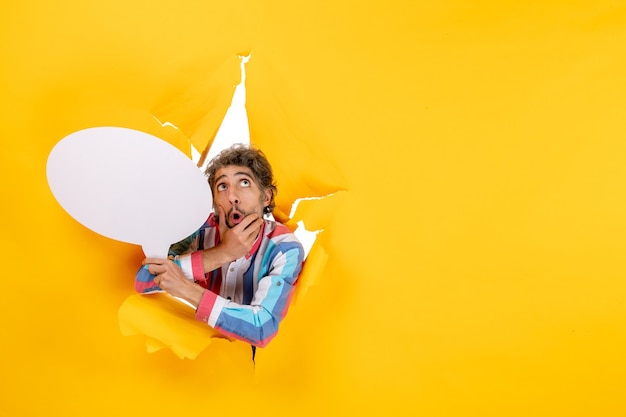 Free photo bewildered young guy holding white balloon and looking up in a torn hole and free background in yellow paper