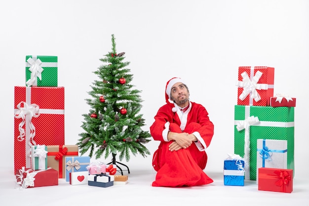 Bewildered young man dressed as Santa claus with gifts and decorated Christmas tree sitting on the ground on white background