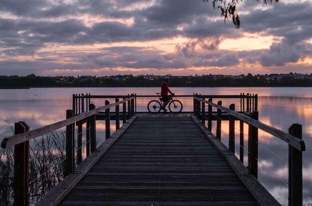 Bicyclist standing on a wooden dock on the water under a cloudy sky during the sunset in the evening