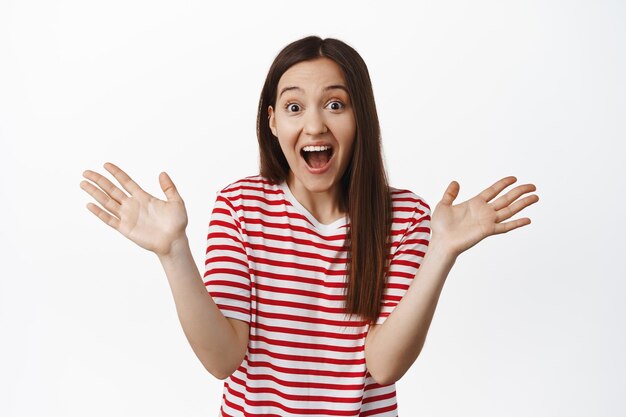 Big announcement. Excited happy 20s woman spread hands sideways, scream from amazement and joy, react to amazing news, standing against white background