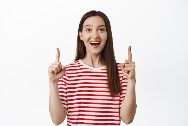 Big announcement on top Excited smiling girl pointing fingers up happy showing sale advertisement logo or banner upwards standing against white background