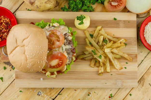 Big burger with some French fries and souse on a wooden tray and potatoes nearby