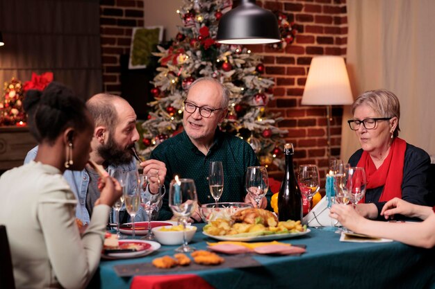 クリスマスを祝い、話し、ホームパーティーのお祝いの夕食の席で伝統的な冬の休日の食べ物を食べる多様な大家族。美しい装飾が施された場所でのクリスマスのお祝い