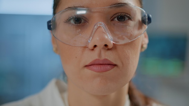 Biochemist with safety goggles and white coat in laboratory, face of scientist looking at camera while she works on chemistry development in lab. Portrait of woman doing research. Close up