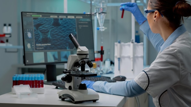 생화학 의사는 책상 위에 현미경을 놓고 전문 의료 실험실에서 컴퓨터를 사용하면서 혈액으로 vacutainer를 분석합니다. 분석 실험을 하는 전문 여성