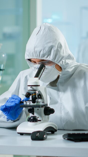 Врач-биолог в комбинезоне проверяет образец ДНК на микроскопе, печатающем на компьютере в оборудованной лаборатории. Изучение эволюции вакцины в медицинской лаборатории с использованием высокотехнологичных и химических инструментов для научных исследований