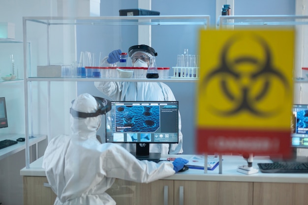 Free photo biologist researchers team wearing protective medical equipment against coronavirus while developing covid19 vaccine working in microbiological hospital laboratory. danger sign on glass