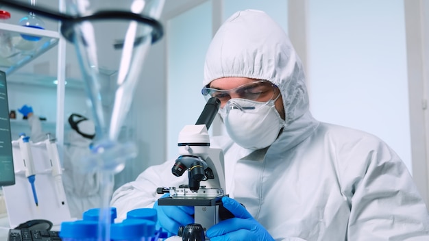 PPE 슈트를 입은 생명공학 과학자는 현미경을 사용하여 실험실에서 DNA를 연구합니다. covid19에 대한 백신 개발의 과학적 연구를 위해 첨단 기술을 사용하여 바이러스 진화를 조사하는 팀