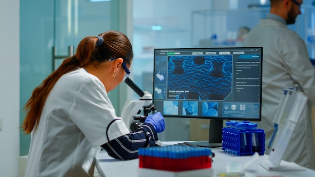 Ученый-биотехнолог изучает лабораторию с помощью микроскопа и печатает на компьютере. Химик изучает эволюцию вируса с помощью высоких технологий для научных исследований разработки вакцины против covid19