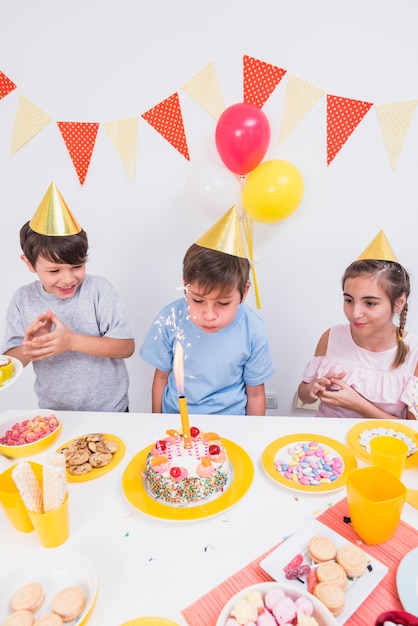 Birthday boy blowing candle with his friends standing behind cake