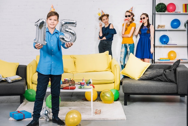 A birthday boy showing numeral 15 foil silver balloons with his friends standing at the background