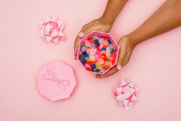 Birthday concept with confetti, bows on pink background flat lay. hands holding gift box.