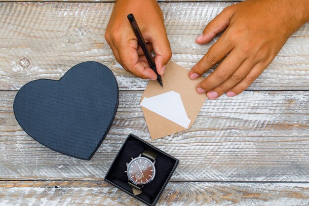 Birthday concept with gift boxes, watch on wooden background flat lay. man writing greeting card with pen.