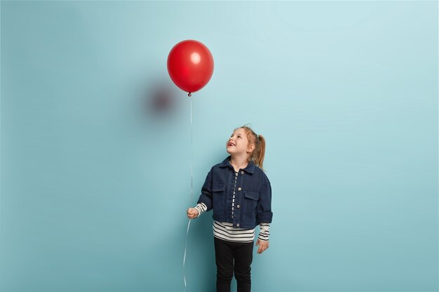 Birthday party and childhood concept. Horizontal shot of glad little child with ginger hair, looks happily upwards on red air balloon, wears fashionable clothes, stands over blue wall