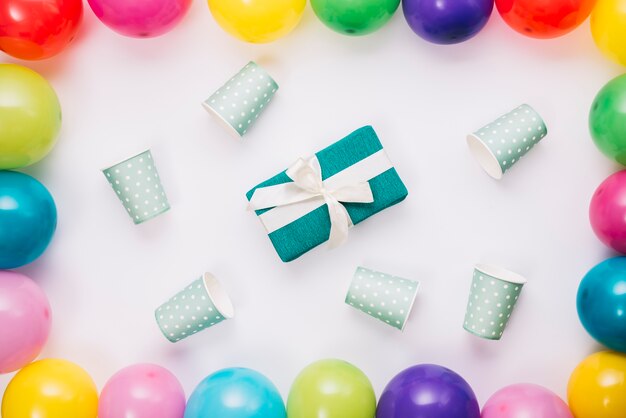 Подарок на день рождения в окружении одноразовой чашки внутри границы воздушных шаров на белом фоне
