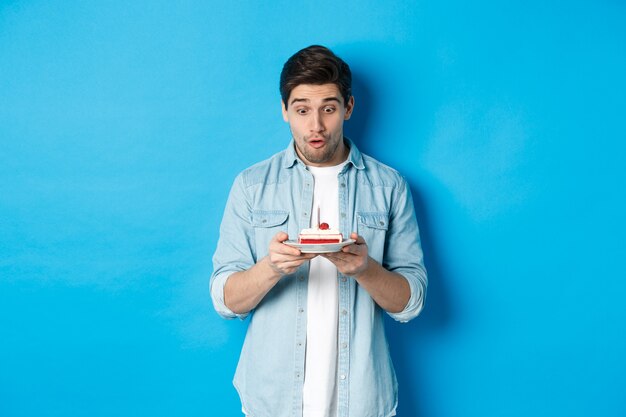 Birthday. Surprised man looking at b-day cake, standing against blue background