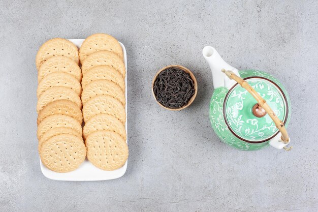 Biscuits lined up on a platter with a teapot and a small bowl of tea leaves on marble surface