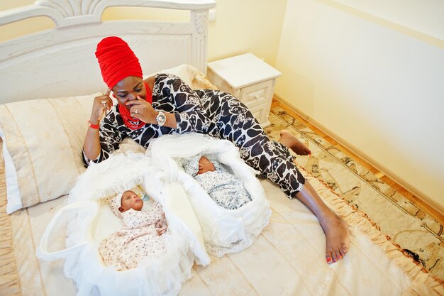 Black african american woman dressed in traditional ethnic clothes laying next to her small babies on the bed