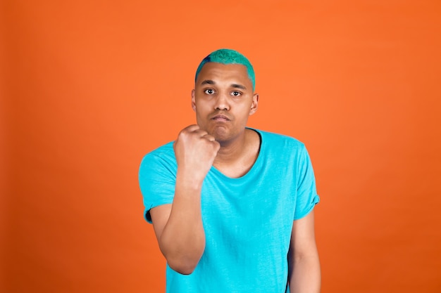 オレンジ色の壁にカジュアルな黒人アフリカ人男性が怒って叫んで拳を上げて不幸で攻撃的な青い髪