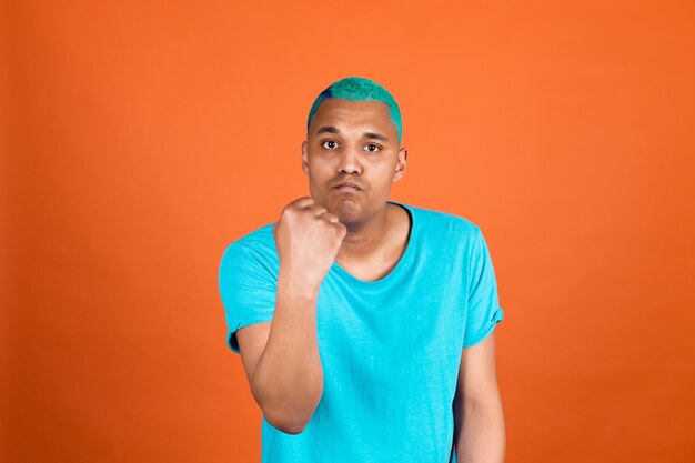 Черный африканский мужчина в повседневной одежде на оранжевой стене с синими волосами кричит сердито, поднимает кулак, несчастный и агрессивный