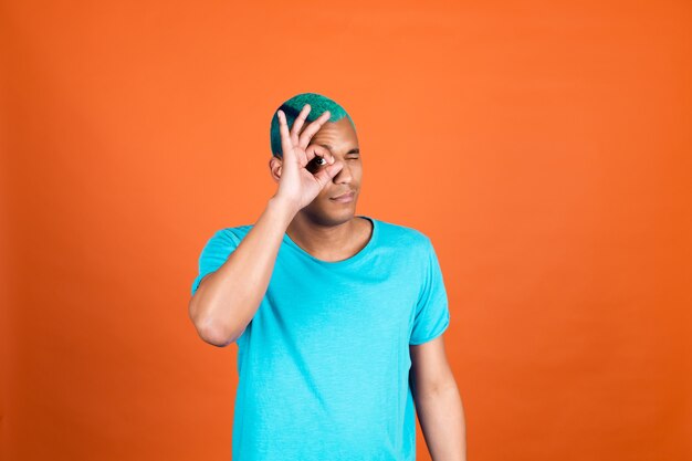 オレンジ色の壁にカジュアルな黒人のアフリカ人男性が片目を覆って大丈夫なジェスチャーを示している青い髪