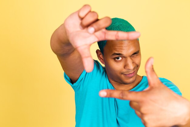 Черный африканский мужчина в повседневной одежде на желтой стене делает фоторамку руками с закрытым глазом и делает снимок