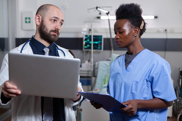병원 병동에서 백인 의사와 함께 일하는 제복을 입은 흑인 아프리카계 미국인 조수, 배경에 있는 아픈 환자, 건강 회복을 위한 현대적인 전문 의료 장비