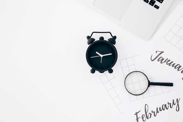 Black alarm clock; laptop and magnifying glasses on paper against white backdrop